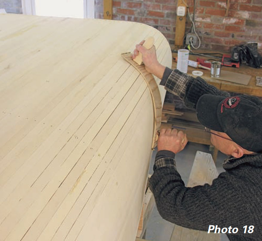 Man uses board sander to sand boat hull at a 45-degree angle to the planking.