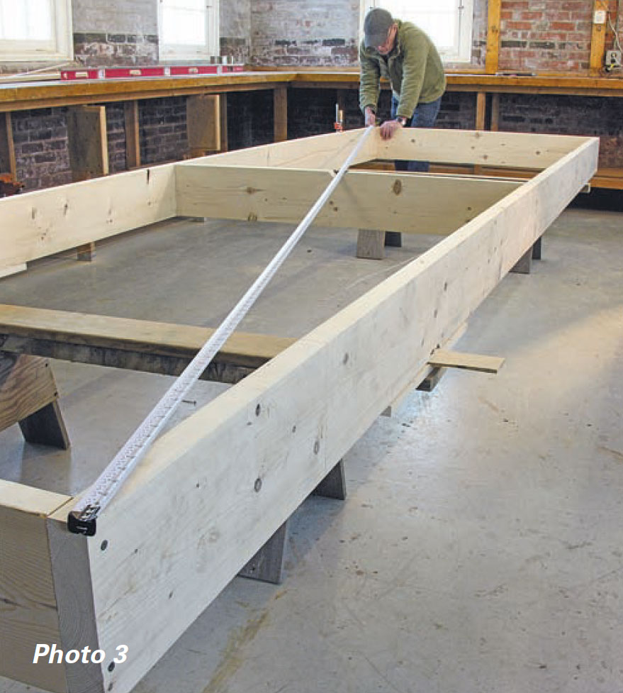 Boatbuilder measures his boat's strongback from corner to corner with measuring tape.