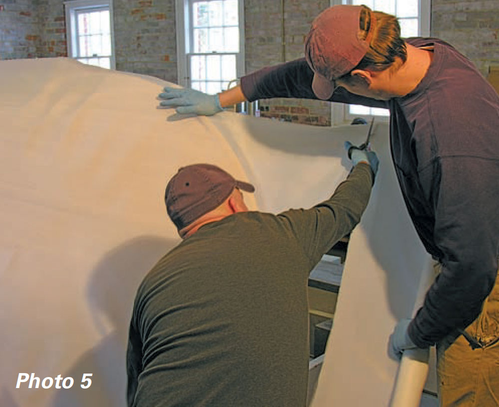 Two men use scissors to cut a piece of fiberglass fabric for a boat.