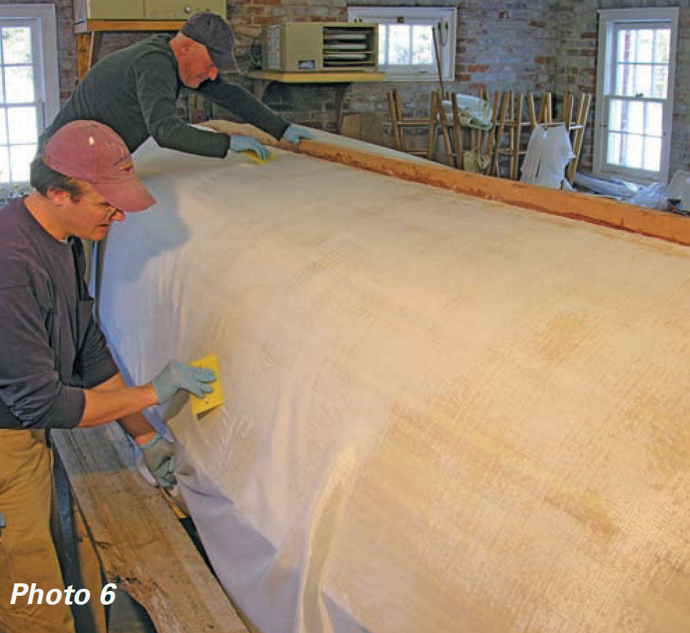 Two boatbuilders use squeegees to smooth out fiberglass cloth on a wooden boat hull.