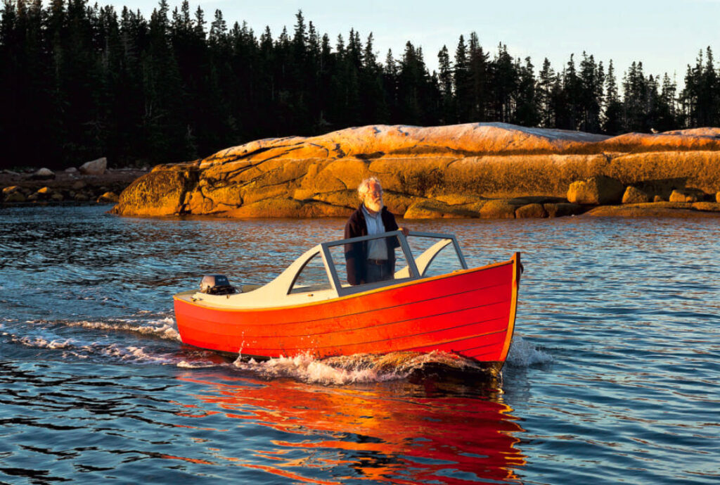 Man stands in a red powerboat on the water with pine trees and a rocky shore in the background.