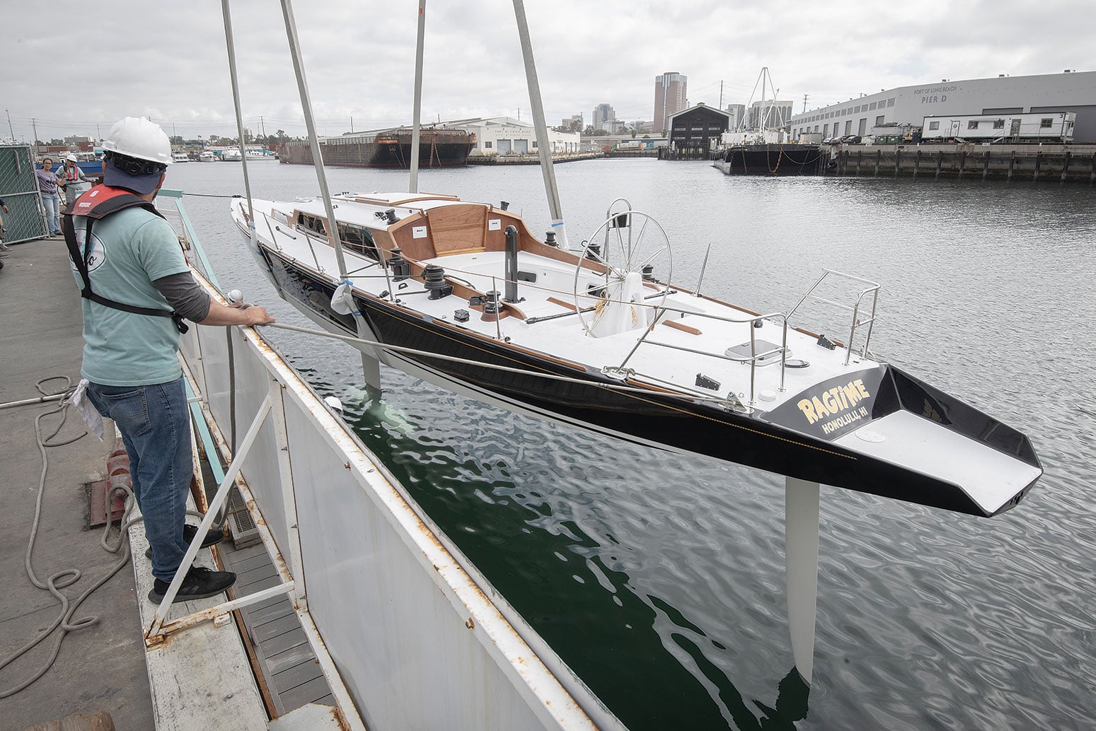 A wooden boat named RAGTIME is lowered into the water by workers.