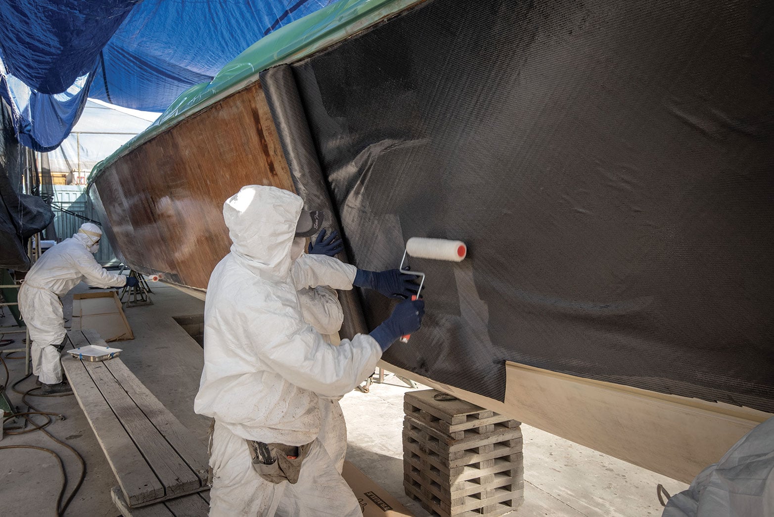 Two workers in PPE apply epoxy to a wooden-boat hull.
