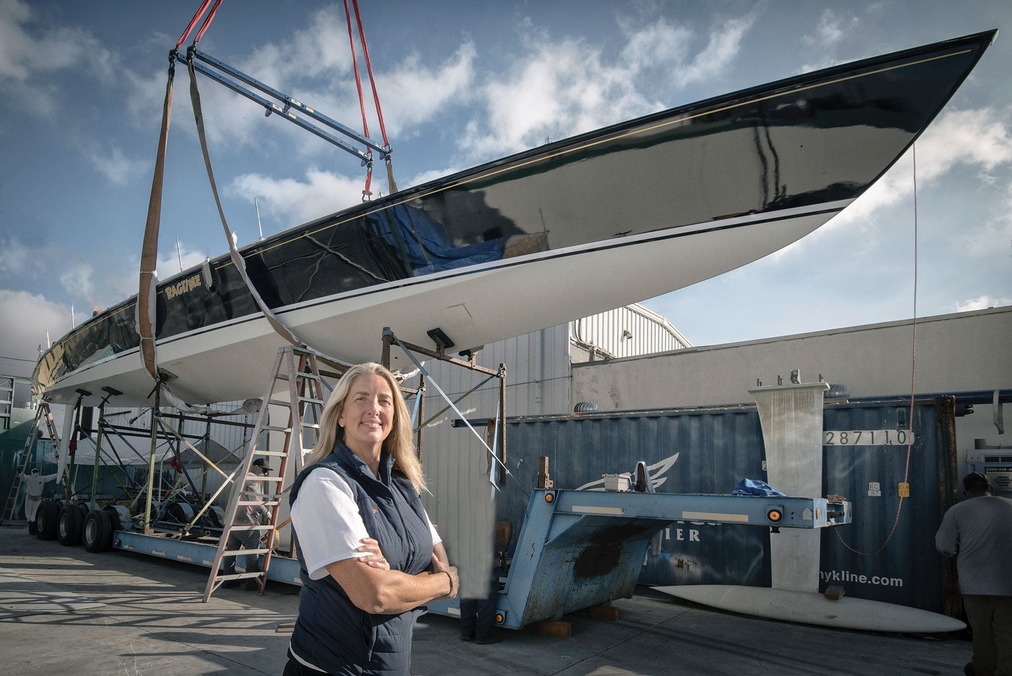 Woman stands in front of a yacht hull suspended by a crane.