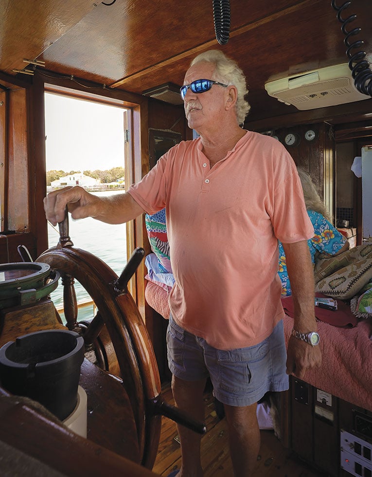 Man in polo shirt and shorts at the wheel of a ship.
