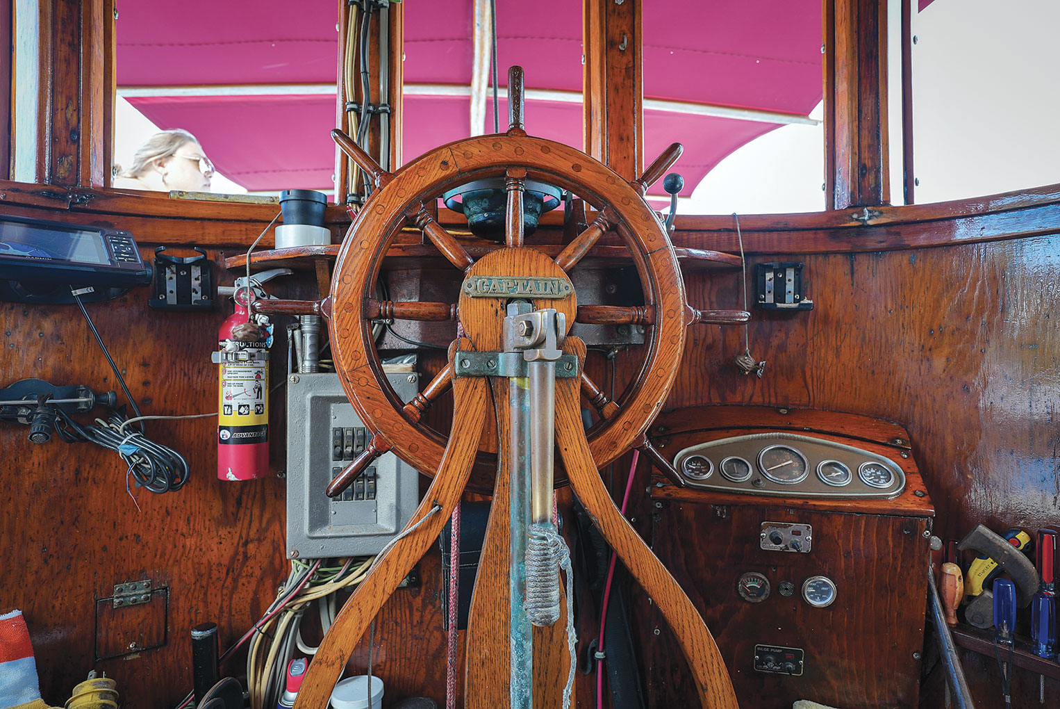 Wooden steering wheel in a ship's pilothouse surrounded by other equipment and a fire extinguisher.