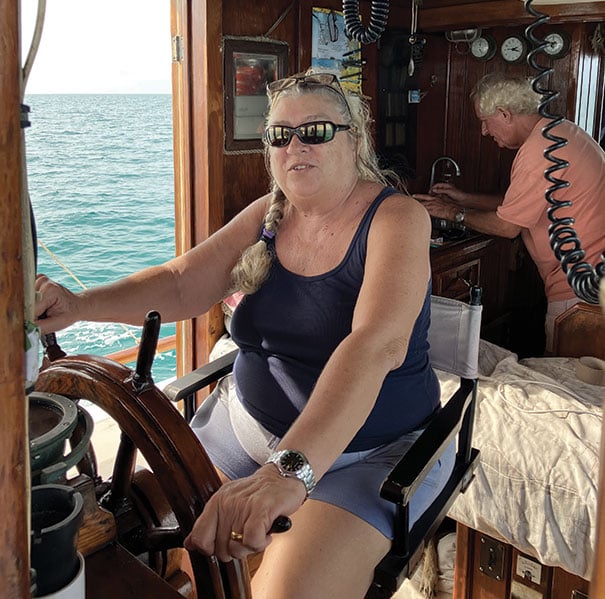 Woman pilots a boat while a man checks a chart in the background.