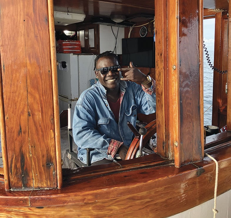Smiling man sits in the pilothouse of a wooden buyboat.